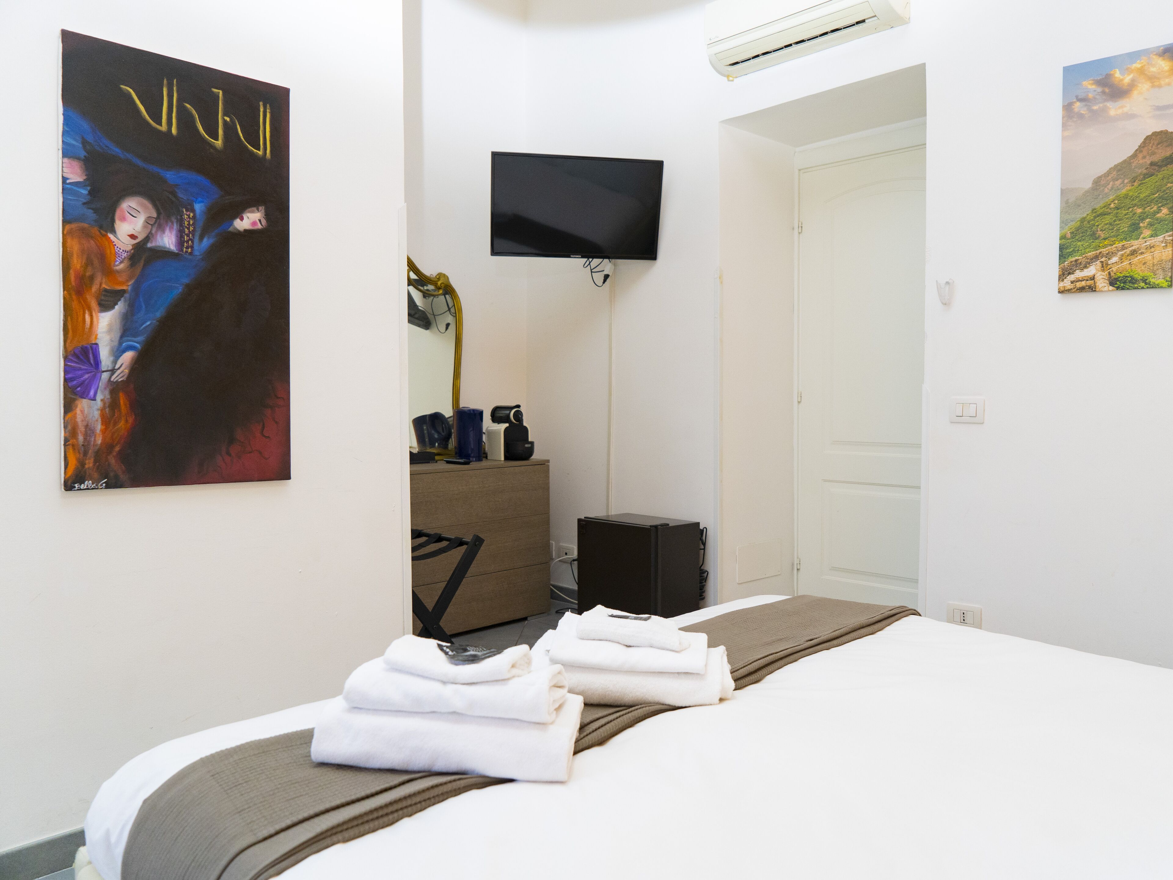 Habitación de hotel con una cama, televisor montado en la pared, un cuadro y una puerta que da a otra habitación.
