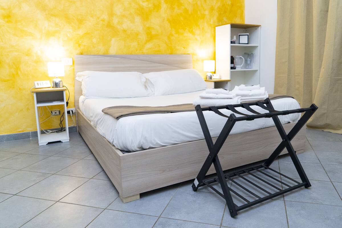 A neatly made bed with white linens, a bedside table with a lamp, and a shelf with toiletries.