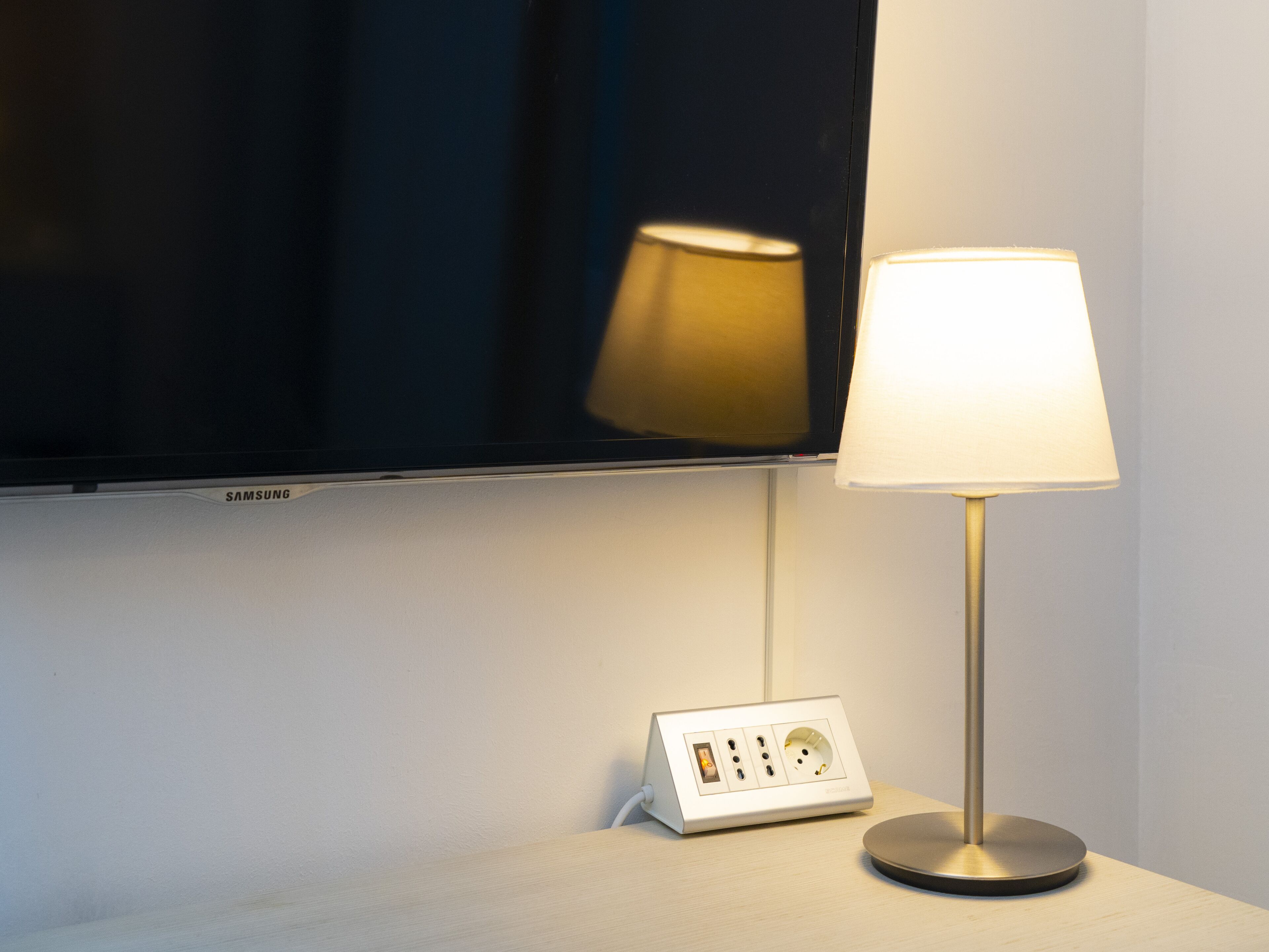 Ein modernes Hotelzimmer mit einem wandmontierten Flachbildfernseher, einem Nachttisch mit Lampe und einer Steckleiste.