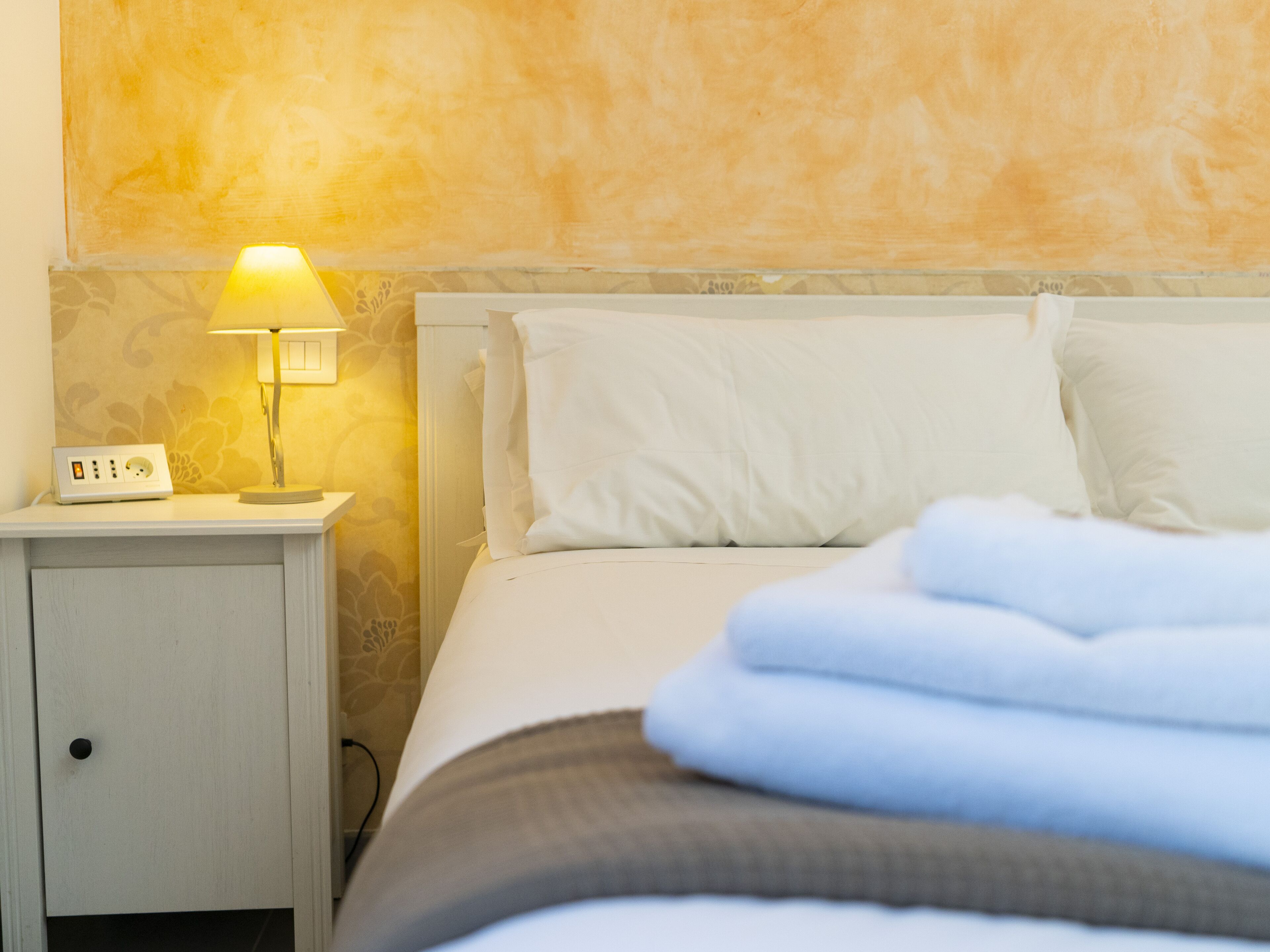 A neatly made bed with white linens and a folded blanket, a bedside lamp, and a power strip on a nightstand.