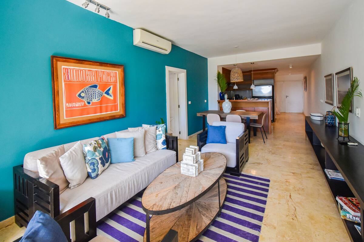 Una sala de estar moderna con una pared de acento color turquesa, un sofá con cojines estampados, una mesa de centro y una zona de cocina al fondo.