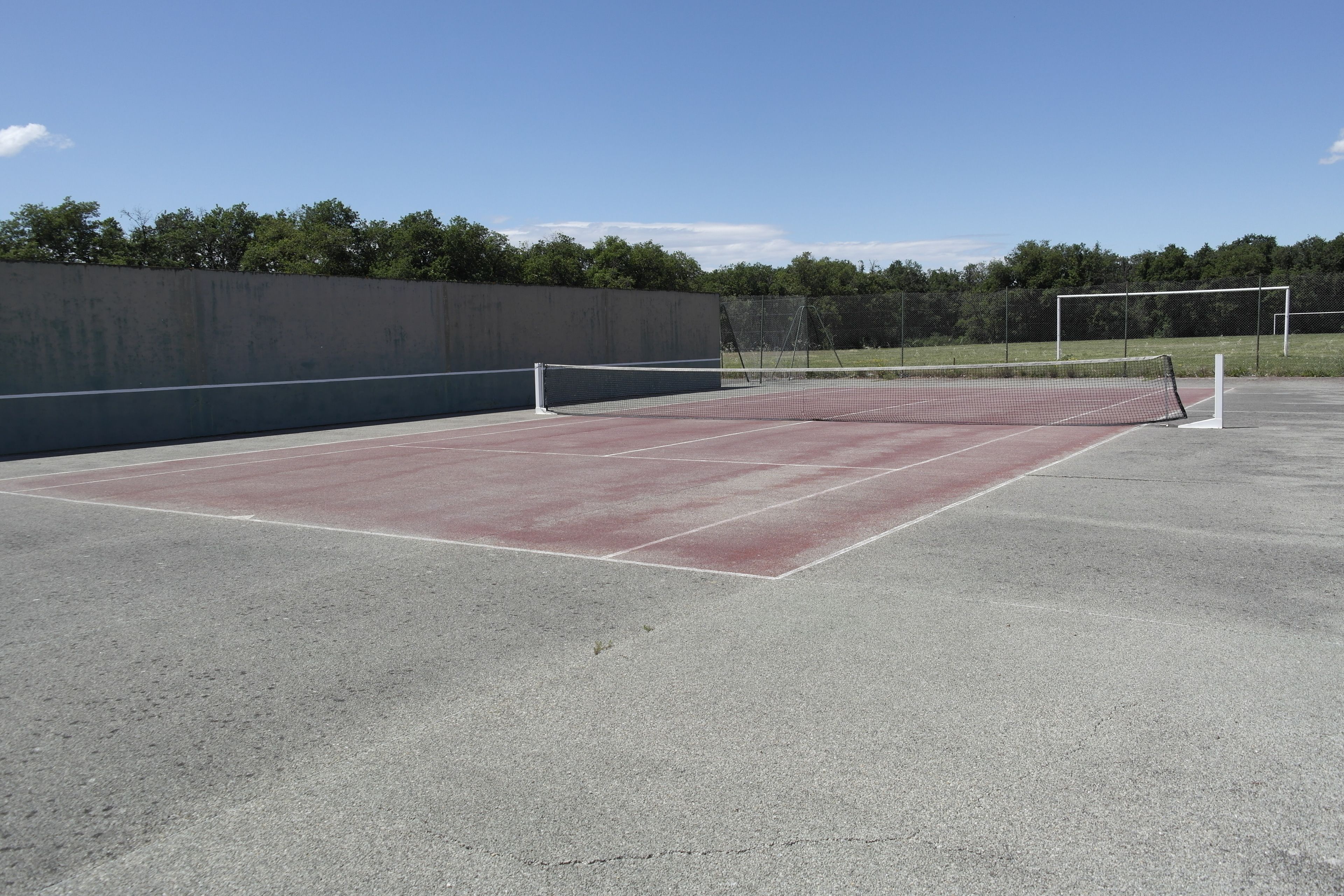An outdoor tennis court with a net, surrounded by a low wall and trees.