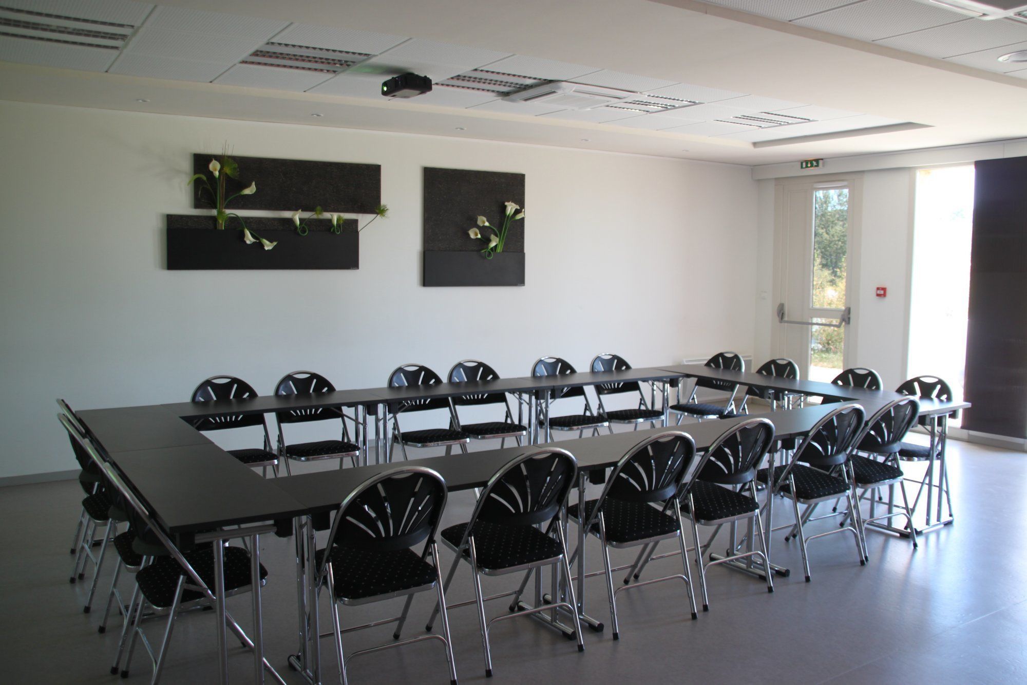 Un salón de conferencias con una mesa larga y sillas, un proyector en el techo y dos arreglos florales en la pared.
