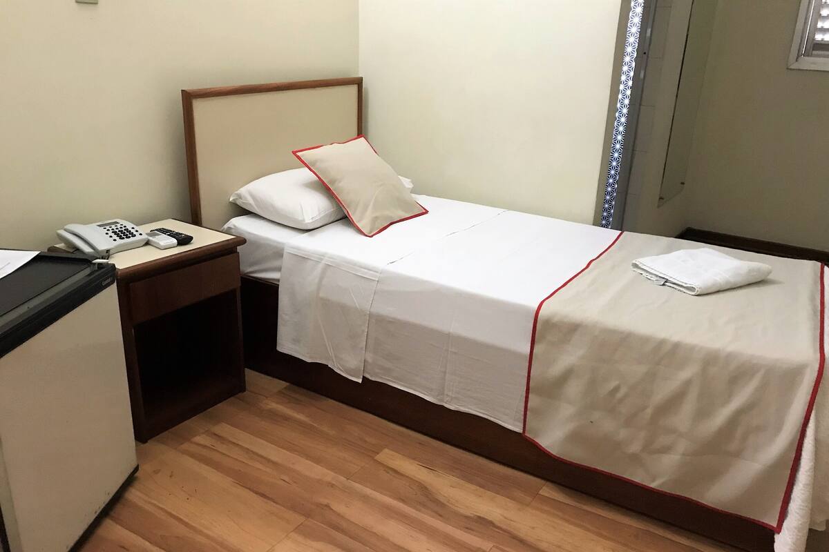 Un lit simple avec une tête de lit en bois, une table de chevet avec un téléphone et une télécommande, et un petit bureau avec un ordinateur.