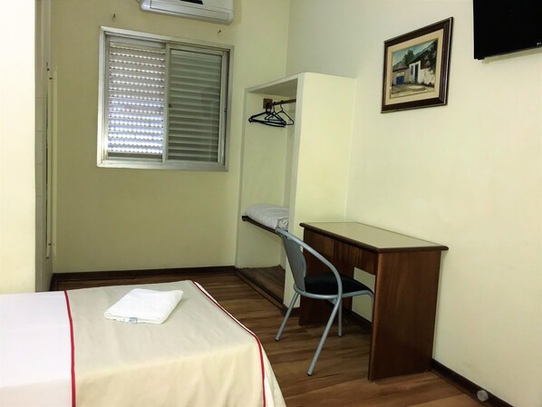 Una habitación pequeña y ordenada con cama, escritorio, silla, ventana con persianas, televisor montado en la pared y un cuadro enmarcado.