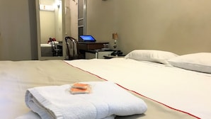 Una cama bien hecha con sábanas blancas y un borde rojo, una computadora portátil sobre una mesita y una silla al fondo.