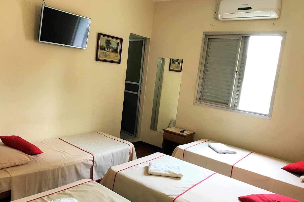 Une chambre avec trois lits simples, une télévision, un climatiseur et une fenêtre avec des stores.