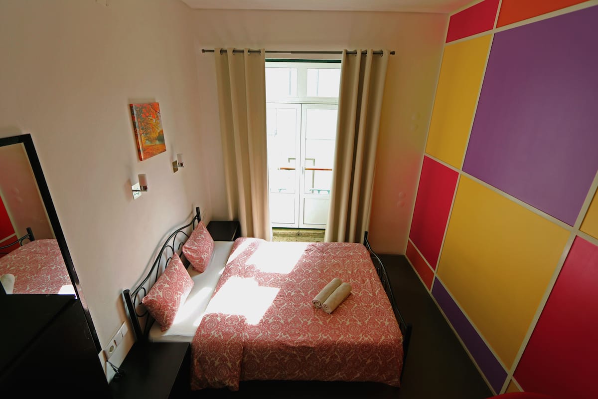 Una habitación con cama, ventana con cortinas, pared con paneles de colores y un cuadro en la pared.