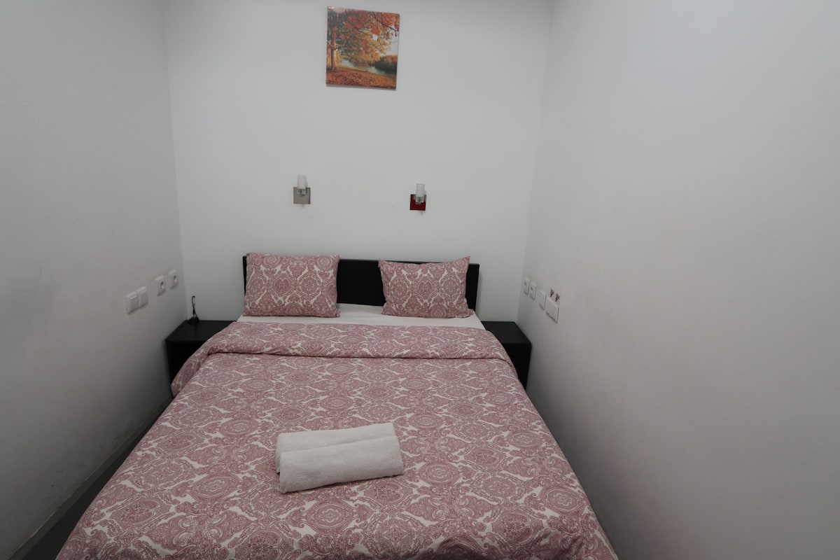 Una cama pequeña y bien hecha con una colcha estampada y almohadas, una mesita de noche con una lámpara, y un aplique en la pared.