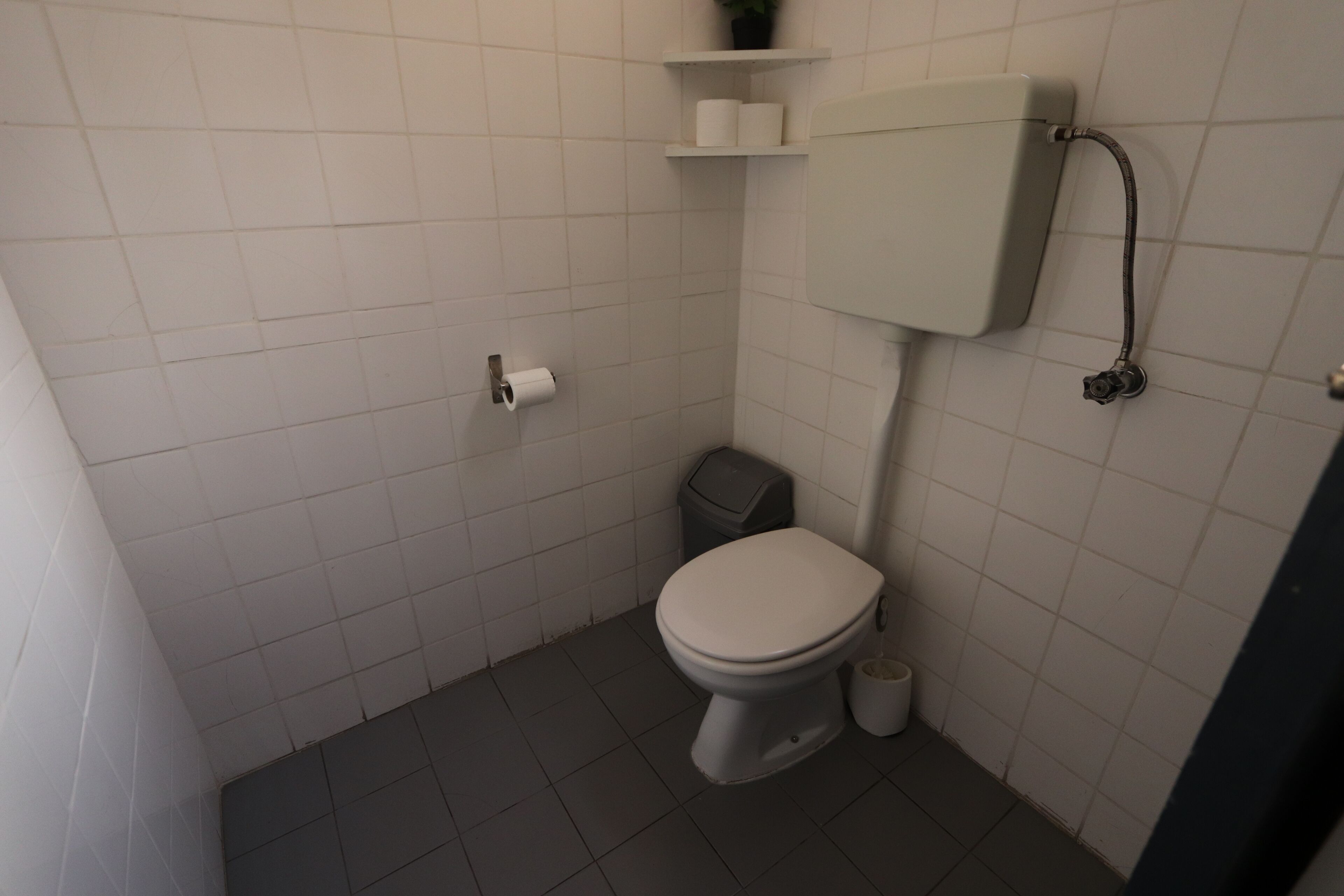 A small, clean bathroom with white tiles, a toilet, a trash bin, and a toilet paper holder.