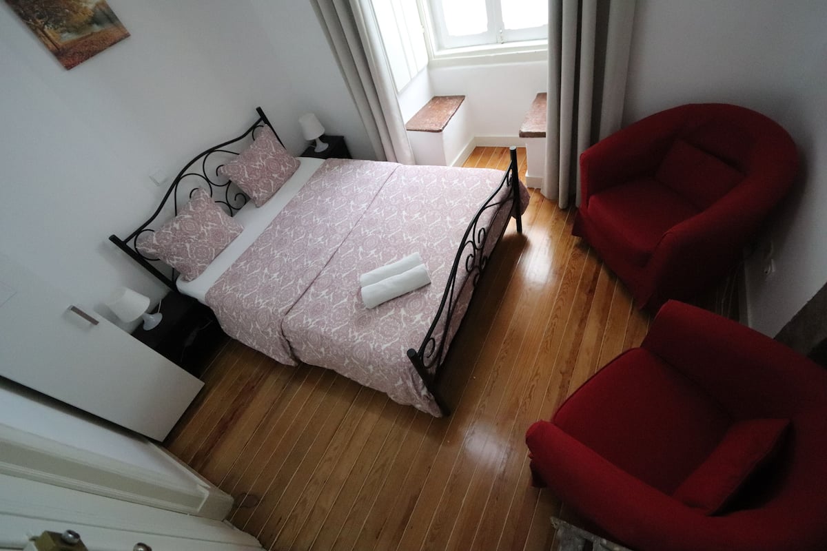 Un dormitorio con una cama, dos sillas rojas, suelo de madera y una ventana con cortinas.