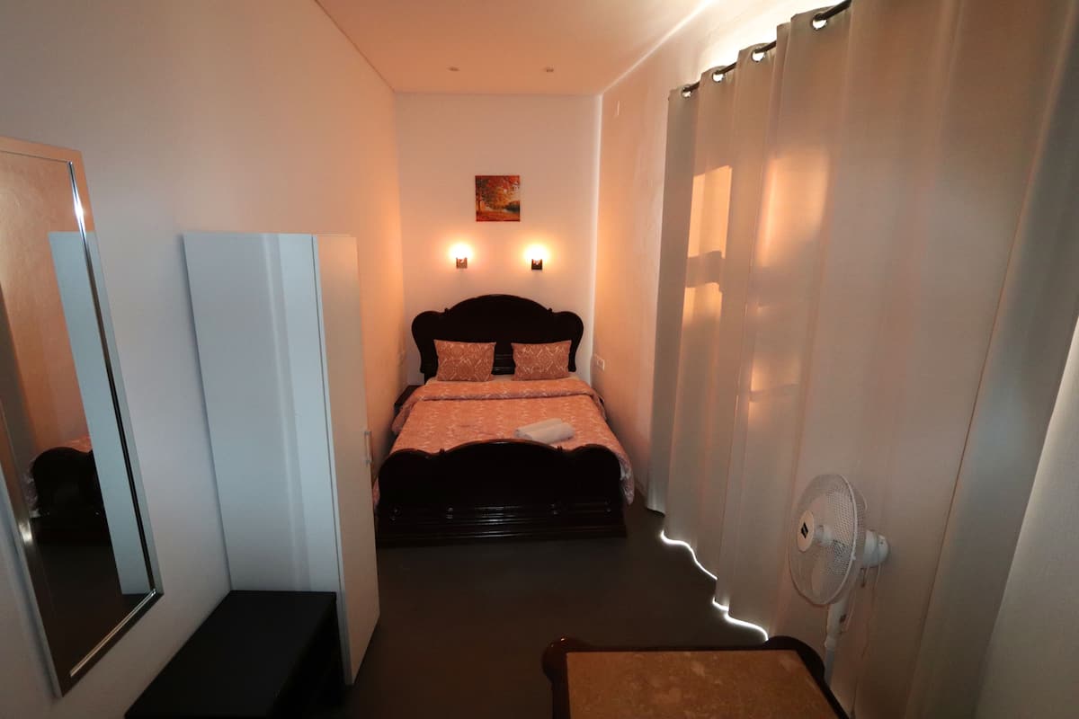 Un dormitorio con cama, mesita de noche, espejo, luz empotrada en la pared y ventilador de techo.