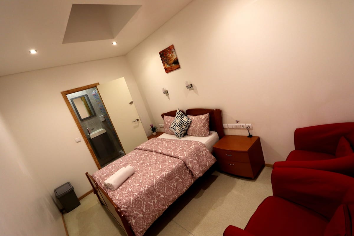 Un dormitorio con una cama, una mesita de noche, una silla roja, una obra de arte colgada en la pared y una puerta que da al baño.