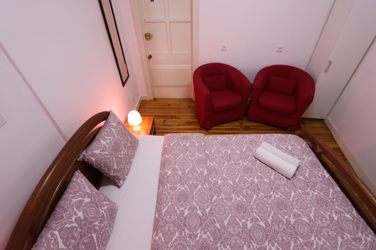Un dormitorio con una cama doble, dos sillones rojos, una mesita de noche con una lámpara y una puerta al fondo.