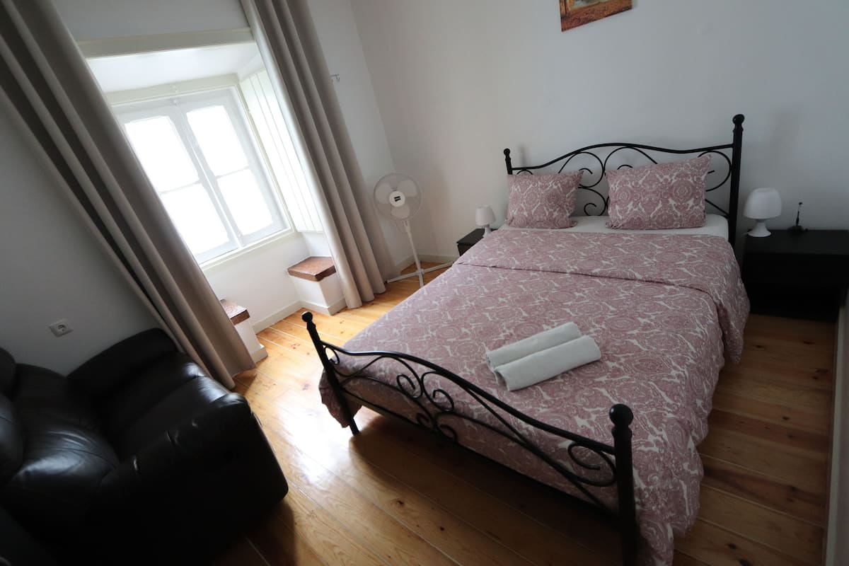 Un dormitorio con una cama, una mesita de noche, una lámpara, un ventilador de pie y una ventana con cortinas.