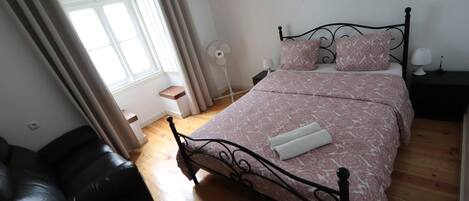 Quarto com cama, mesa de cabeceira, abajur, ventilador e janela com cortinas.