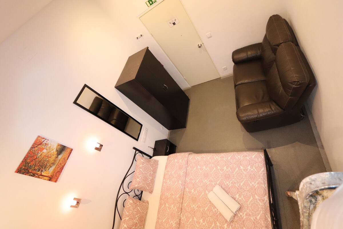 Habitación pequeña con una cama, un sillón de cuero marrón, una mesita y un televisor montado en la pared.