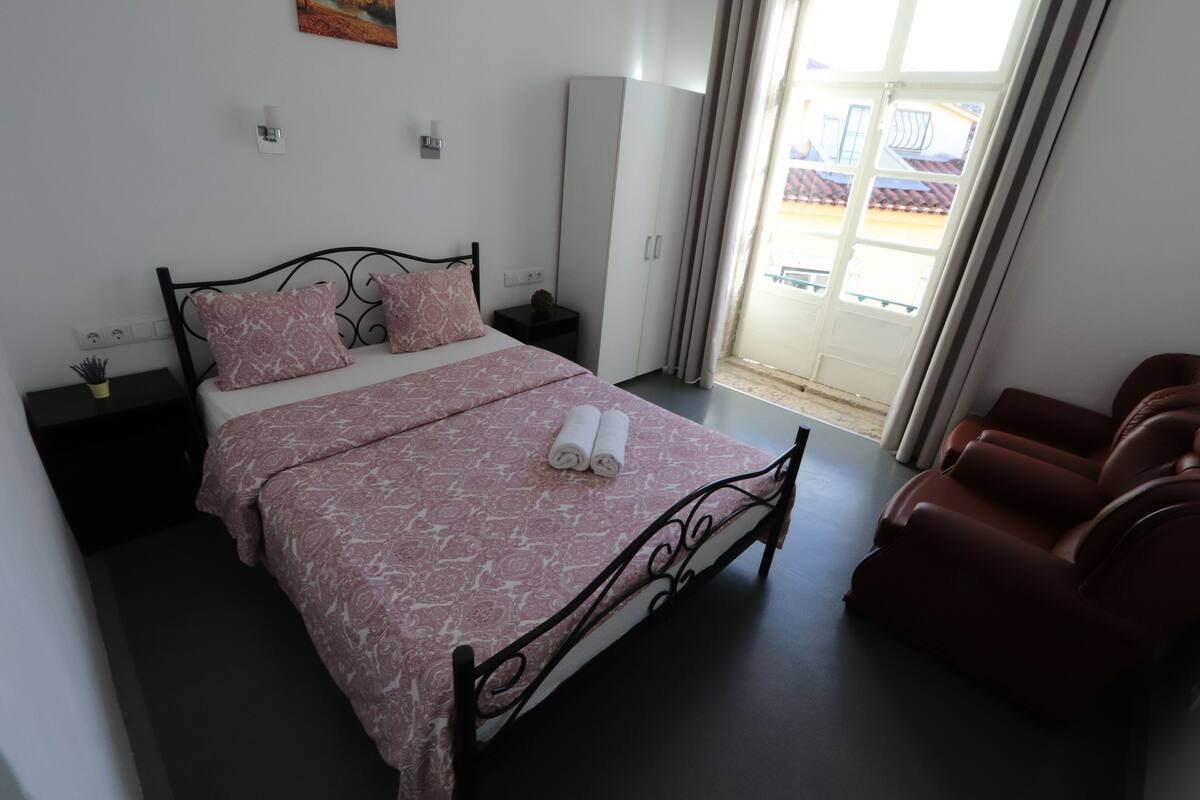 Un dormitorio con cama, armario, zona de estar y vistas por la ventana.