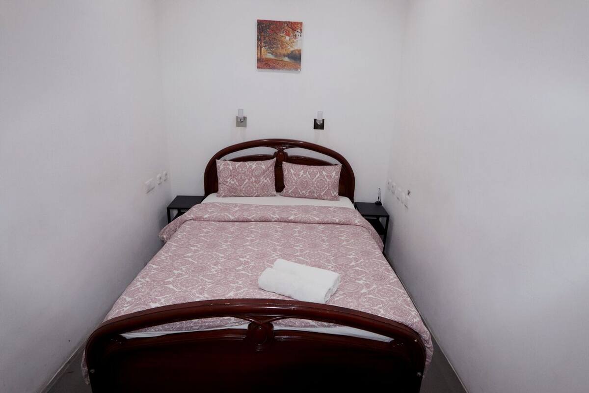 Una cama individual con estructura de madera y una colcha estampada, una toalla blanca sobre la cama y un cuadro pequeño en la pared.