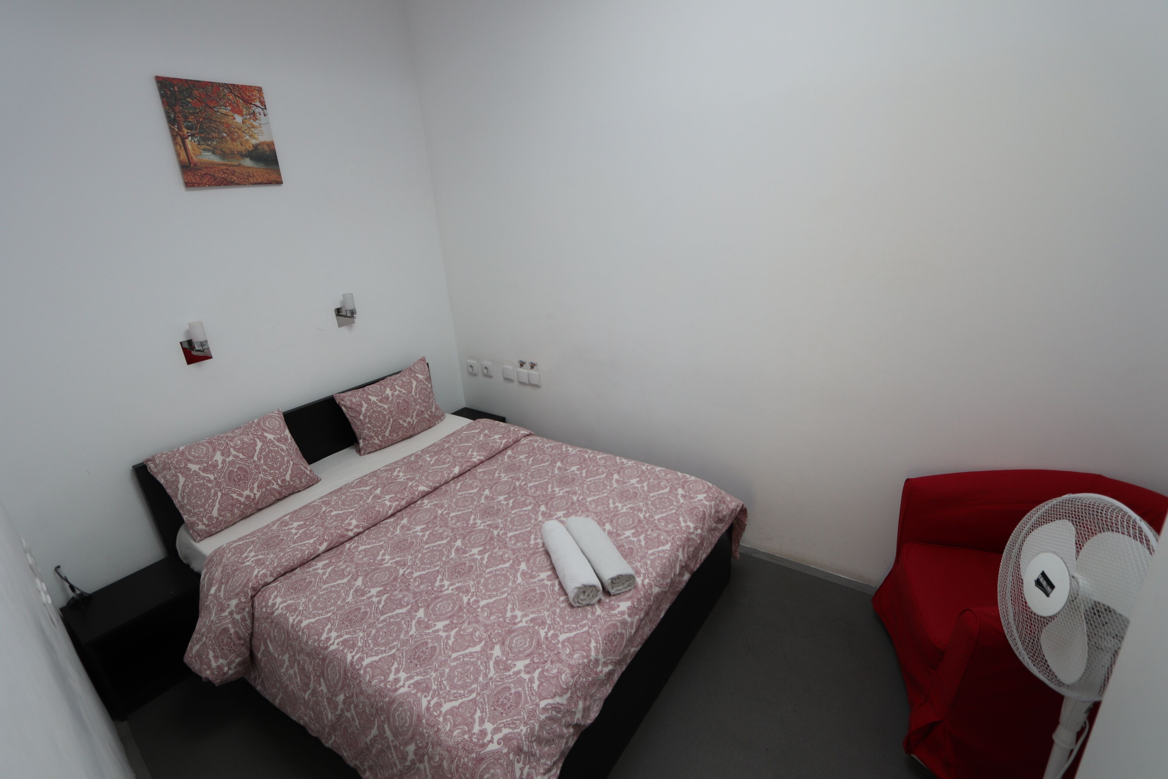 Quarto com cama, almofadas, uma cadeira vermelha, um ventilador e um quadro na parede.