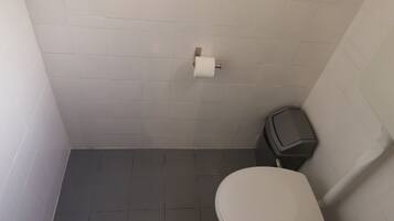 Une petite salle de bain avec des toilettes blanches, une poubelle et un porte-rouleau de papier toilette fixé au mur.
