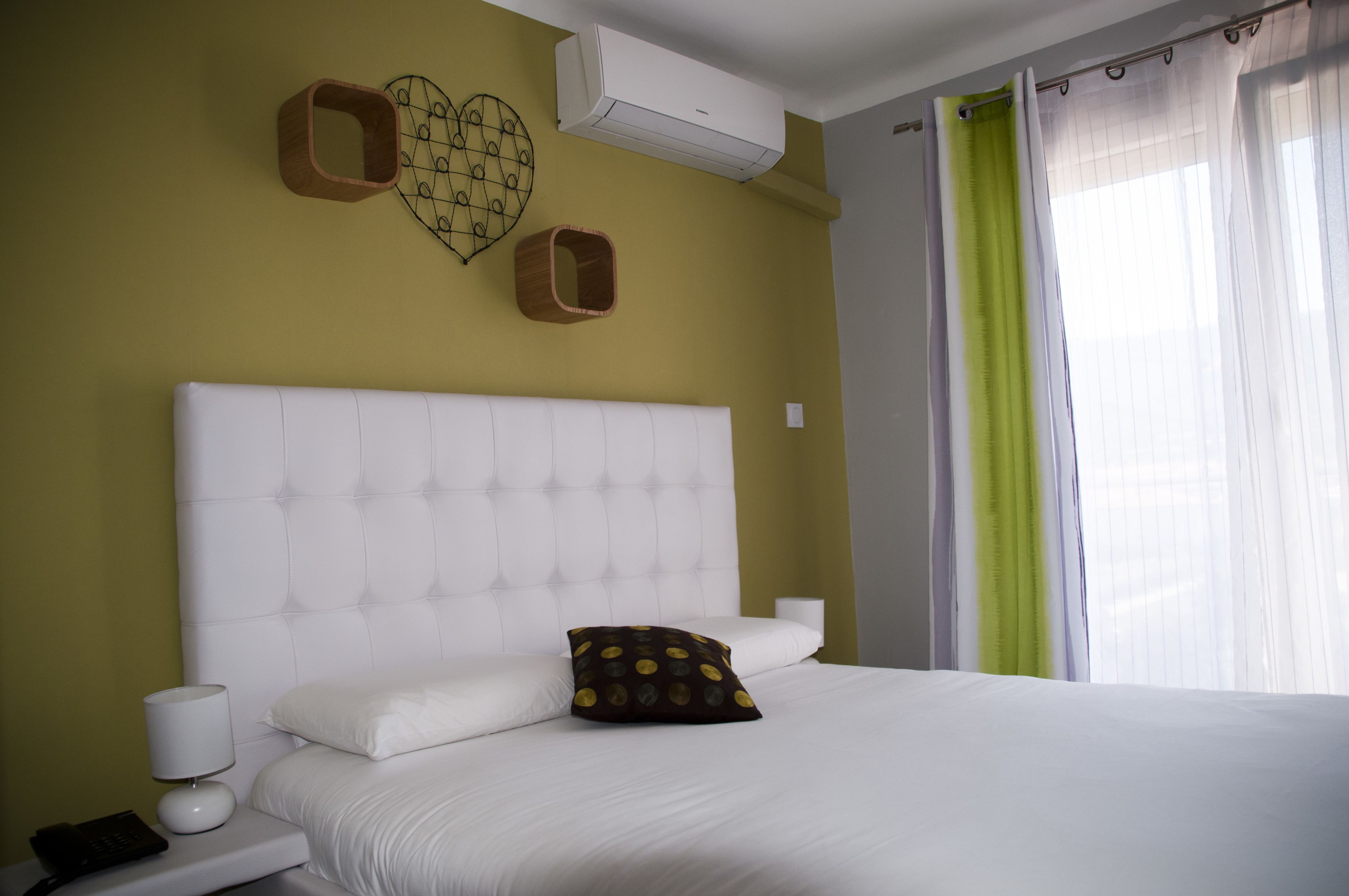 Een opgemaakt bed met een wit hoofdeinde, een nachtkastje met een lamp, en een wandairconditioner.