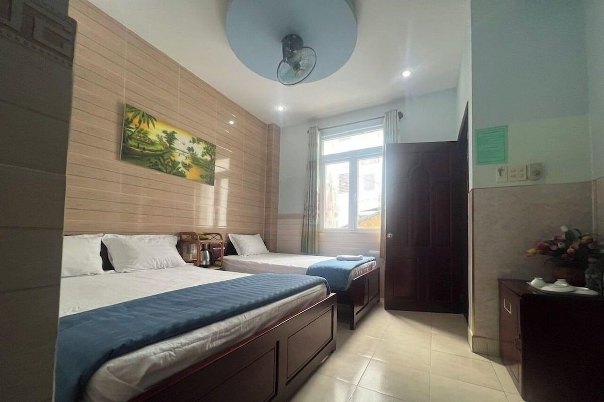 A hotel room with two beds, a ceiling fan, a window with curtains, and a painting on the wall.
