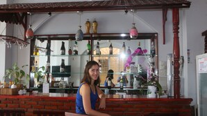 Una mujer sentada en un bar con mostrador de ladrillo, estantes con botellas y luces colgantes.