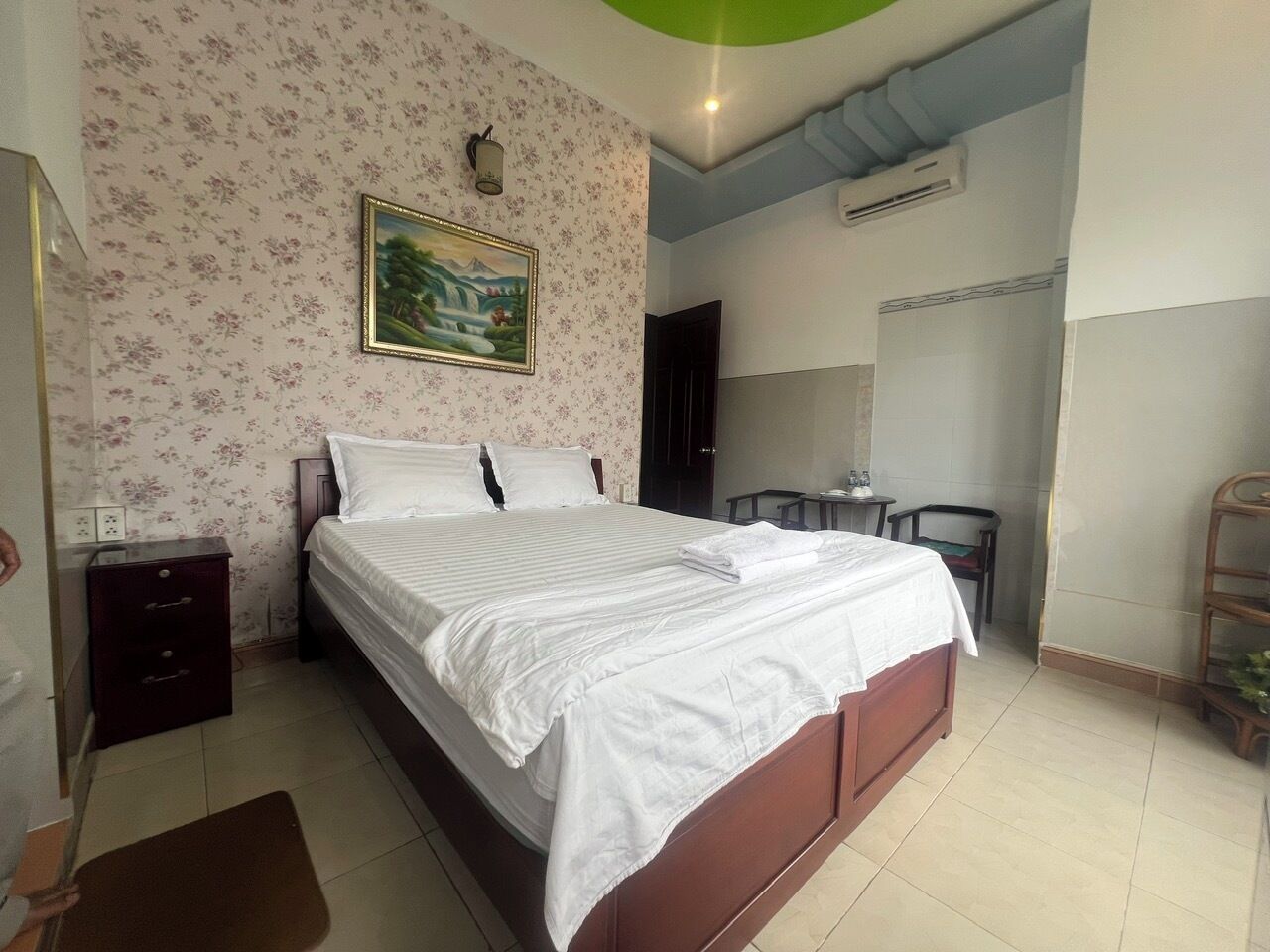 A neatly made bed in a room with floral wallpaper, a small bedside table, and a chair.