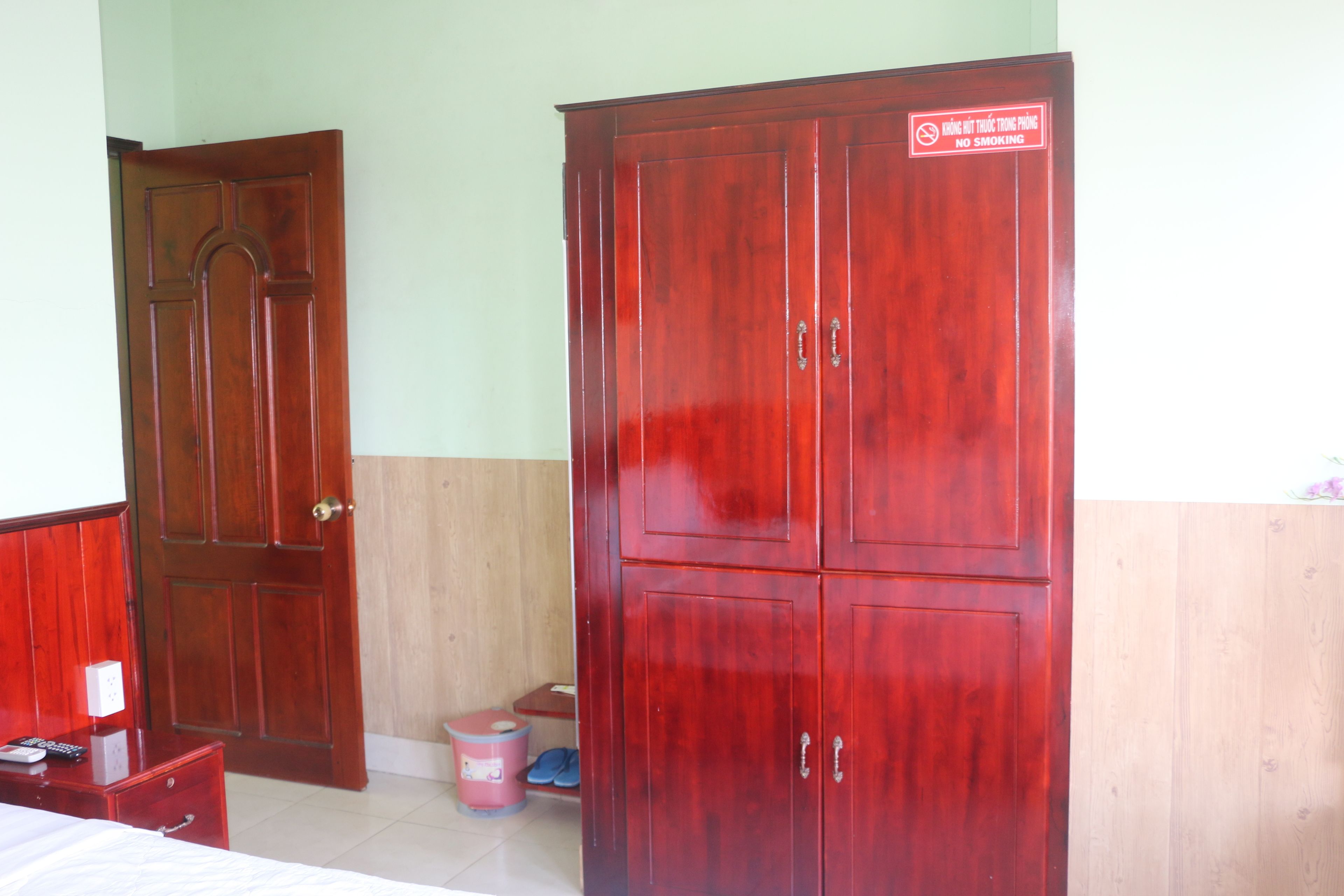 A room with a wooden wardrobe, a bed, a dresser, and a trash bin.