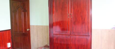 A room with a wooden wardrobe, a bed, a dresser, and a trash bin.