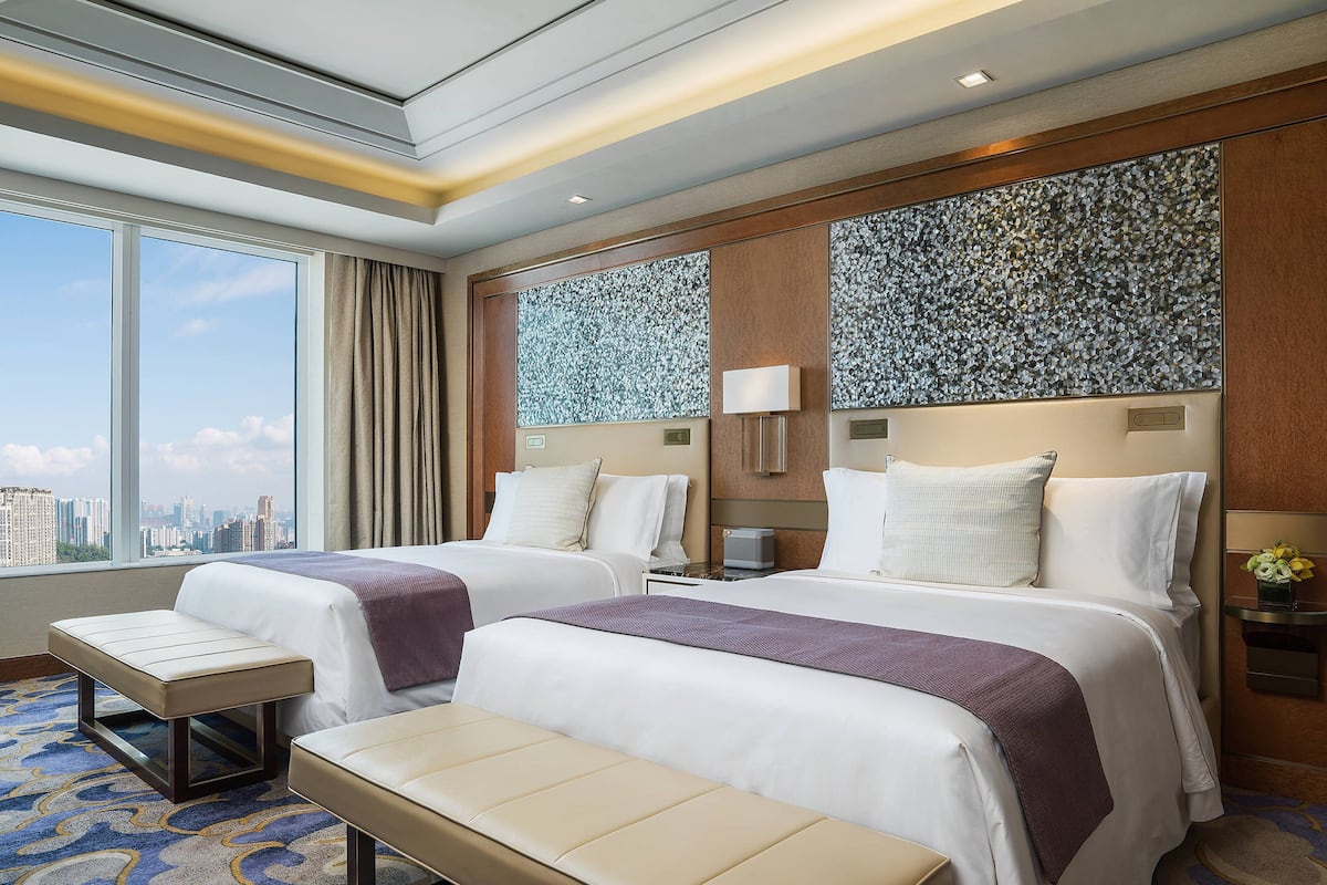 A hotel room with two beds, a bench, a large window with a city view, and a decorative wall panel.