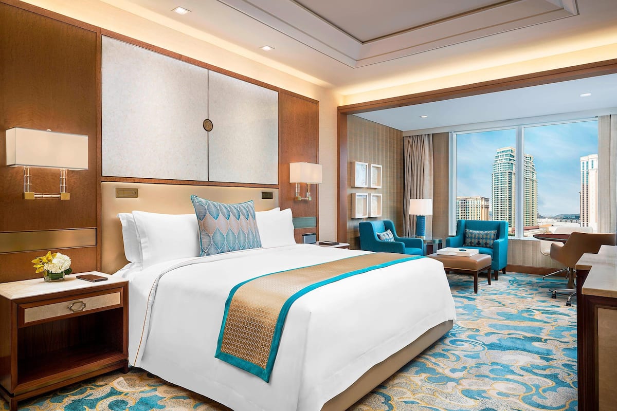 A hotel room with a large bed, a desk, a chair, and a view of tall buildings.