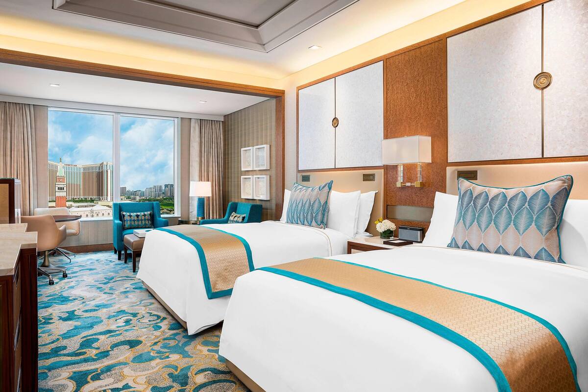 A hotel room with two beds, a desk, and a view of a cityscape.