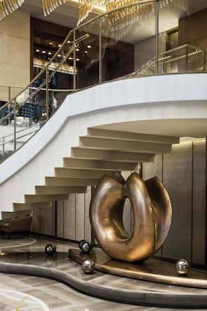 A modern hotel interior with a curved staircase, a large metallic sculpture, and a view of the cityscape through the glass railings.