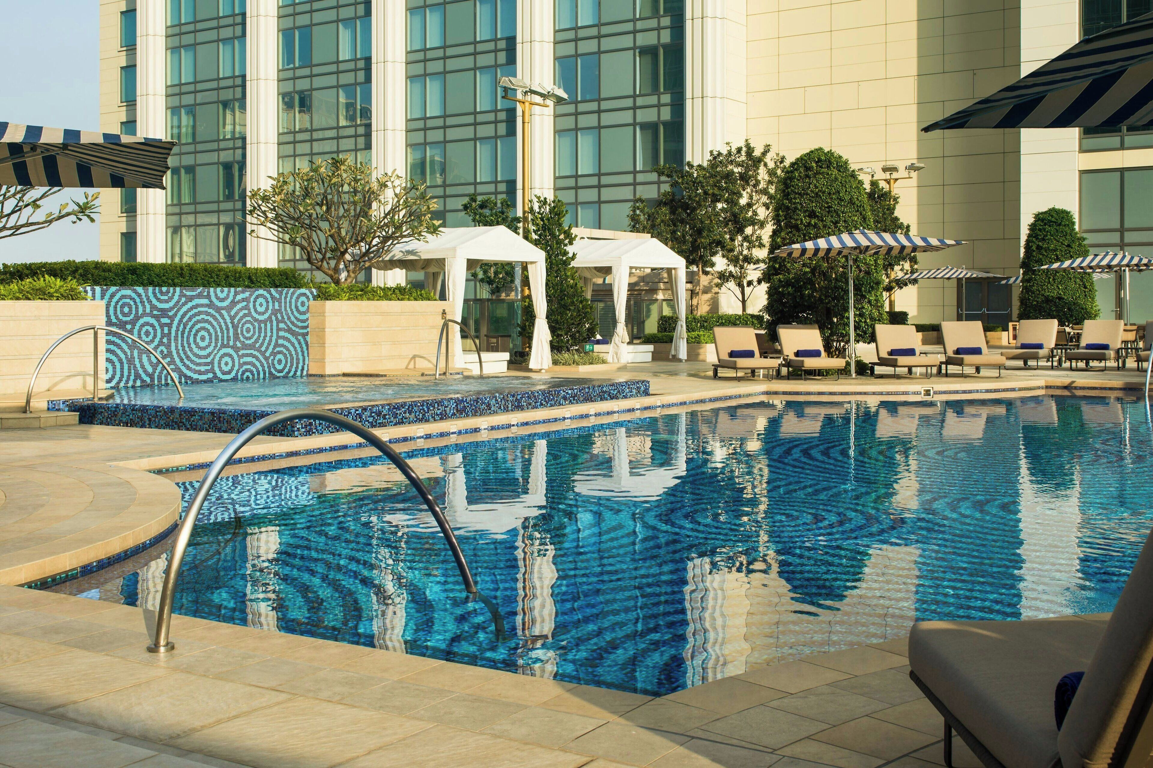 A pool area with lounge chairs and umbrellas, a white tent, and a building with large windows in the background.