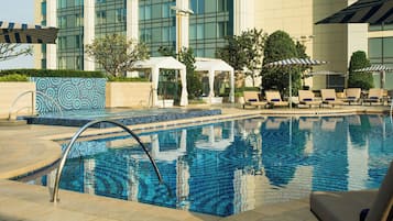 A pool area with lounge chairs and umbrellas, a white tent, and a building with large windows in the background.
