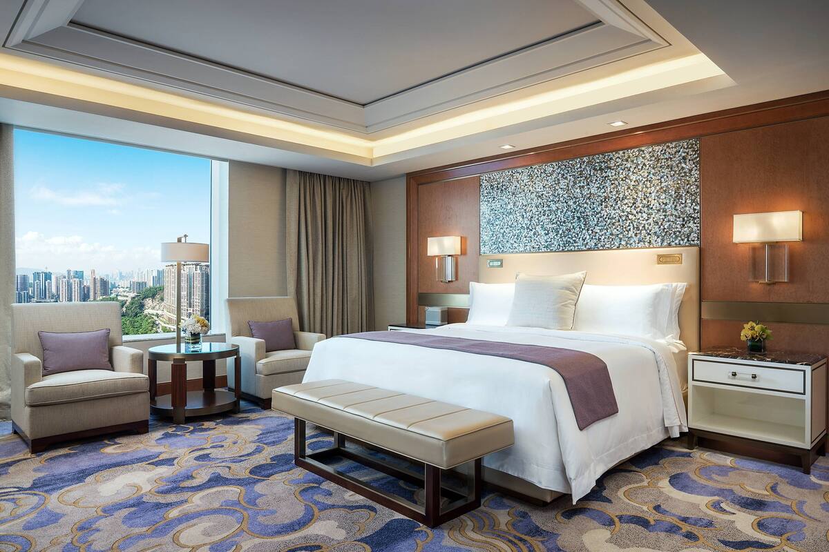 A hotel room with a large bed, two armchairs, a bench, a small table, and a nightstand.