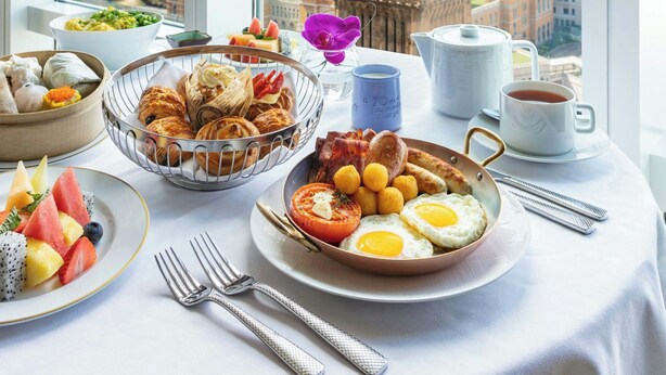 Una mesa de desayuno con diversos platos, vista de un paisaje urbano con una torre de reloj y una ventana con persianas.