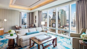 Habitación de hotel con un ventanal grande, un sofá, una mesita de café y vistas a un paisaje urbano que incluye una torre del reloj.