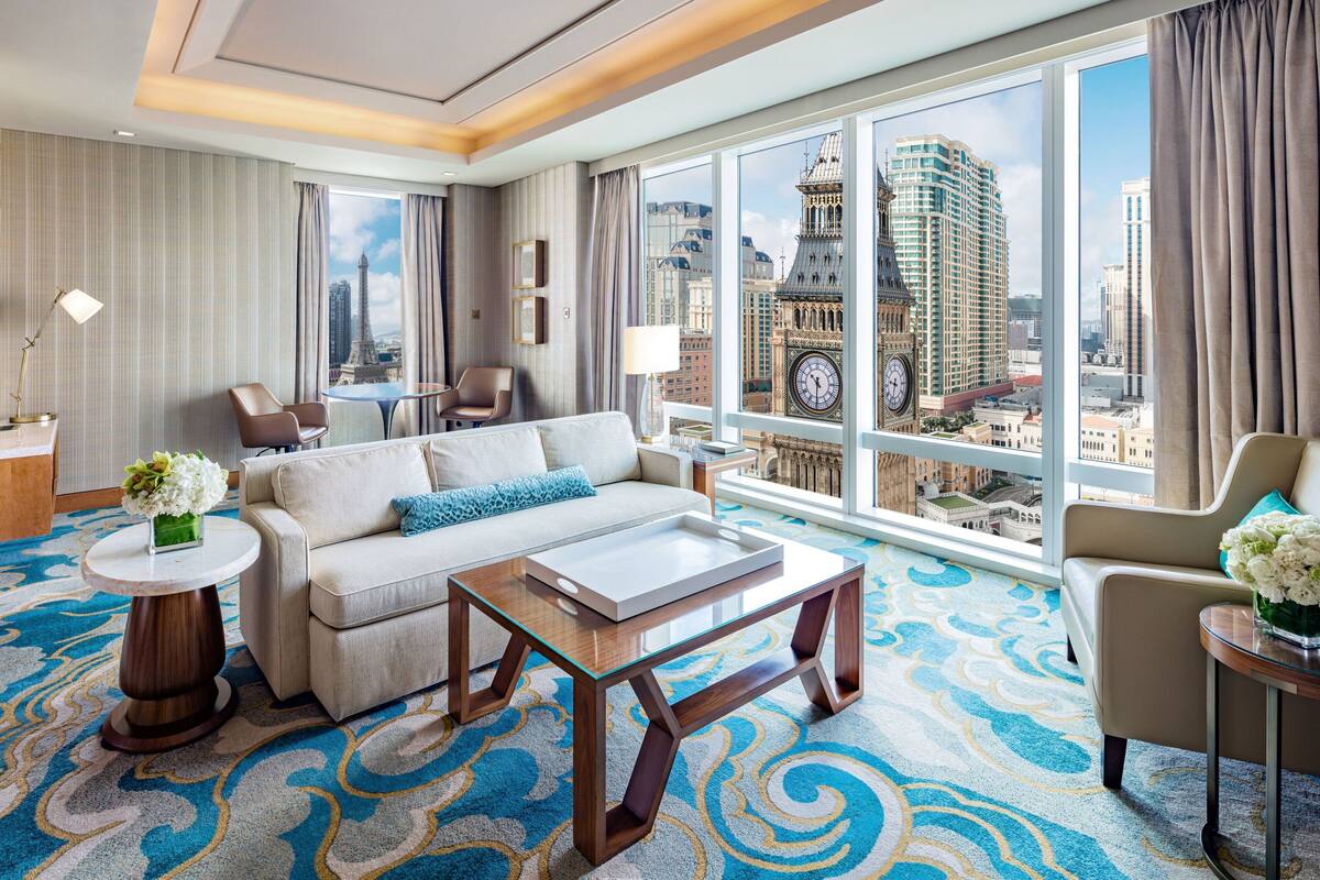 A hotel room with a large window, a sofa, a coffee table, and a view of a cityscape including a clock tower.
