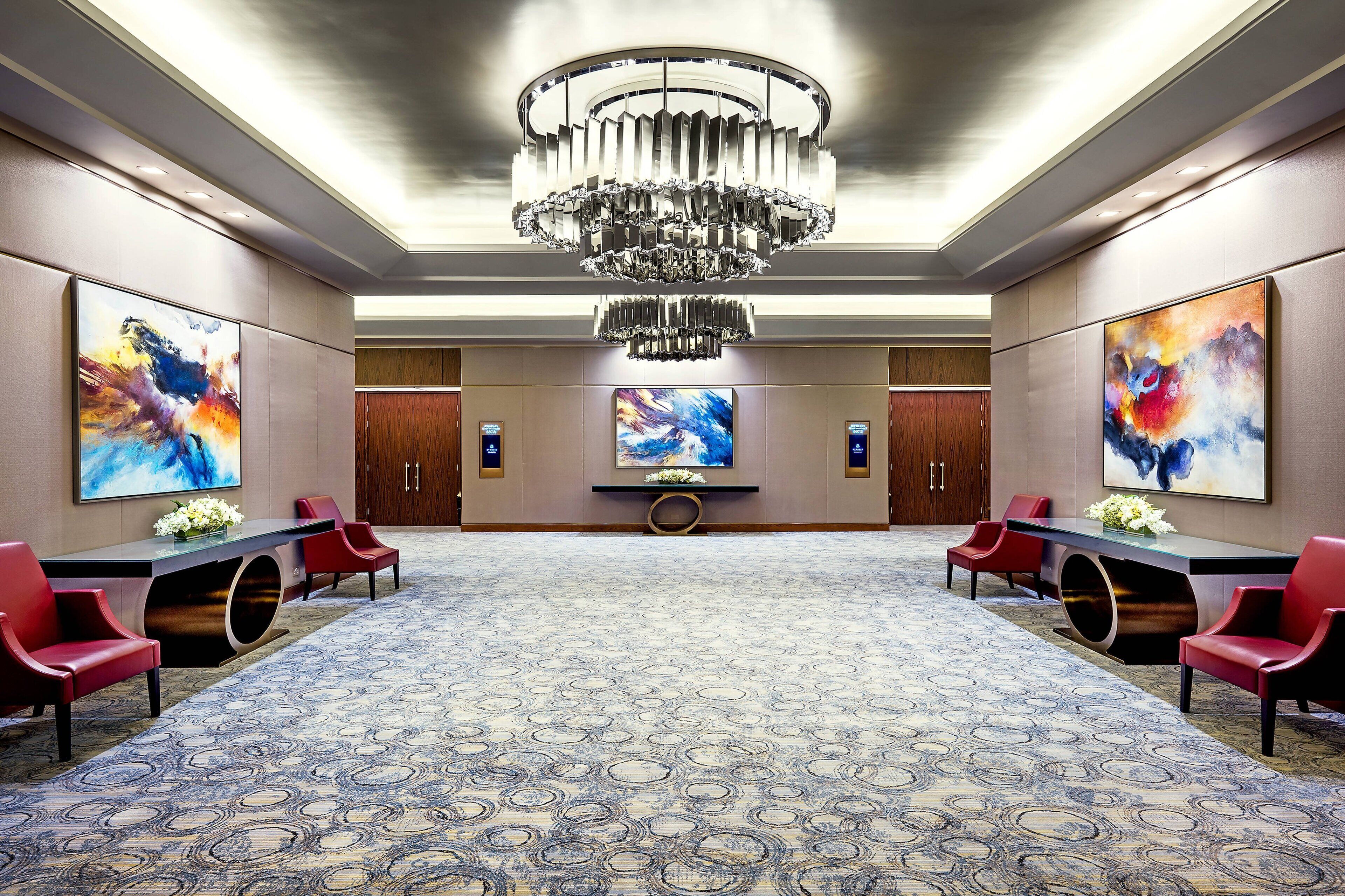 A spacious conference room with a large chandelier, two side tables with red chairs, and abstract paintings on the walls.