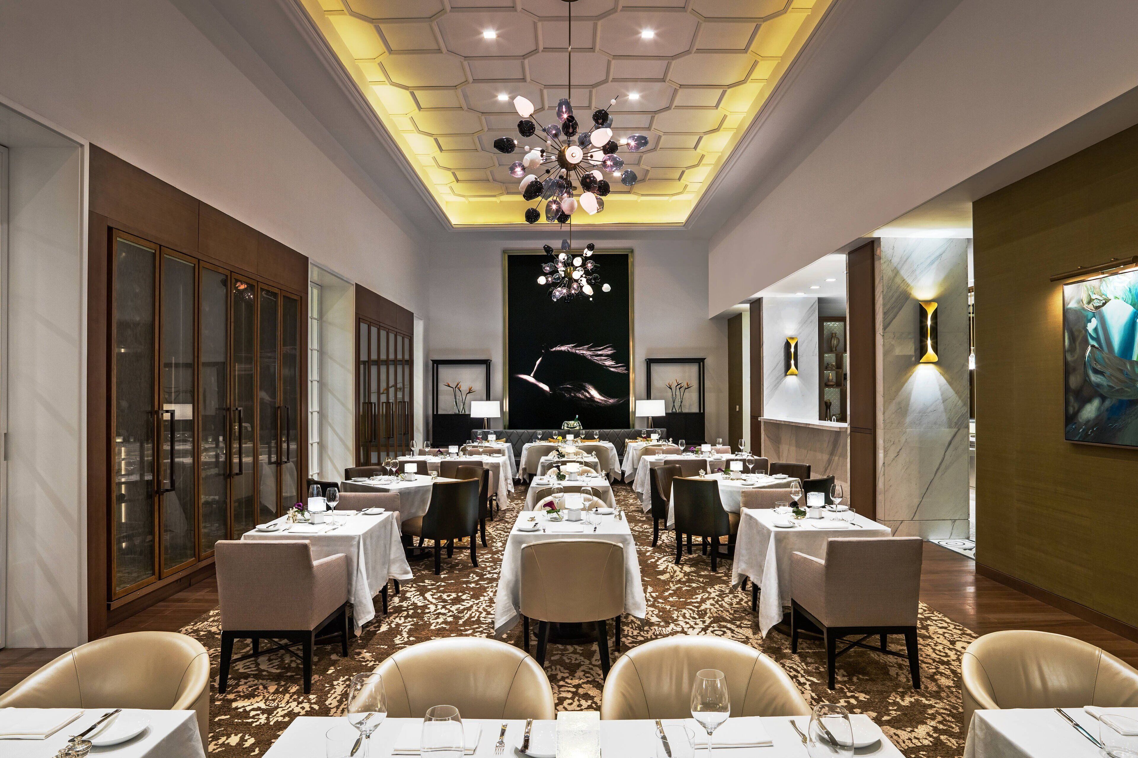 A spacious dining area with multiple tables and chairs, a decorative ceiling, and a large painting on the wall.