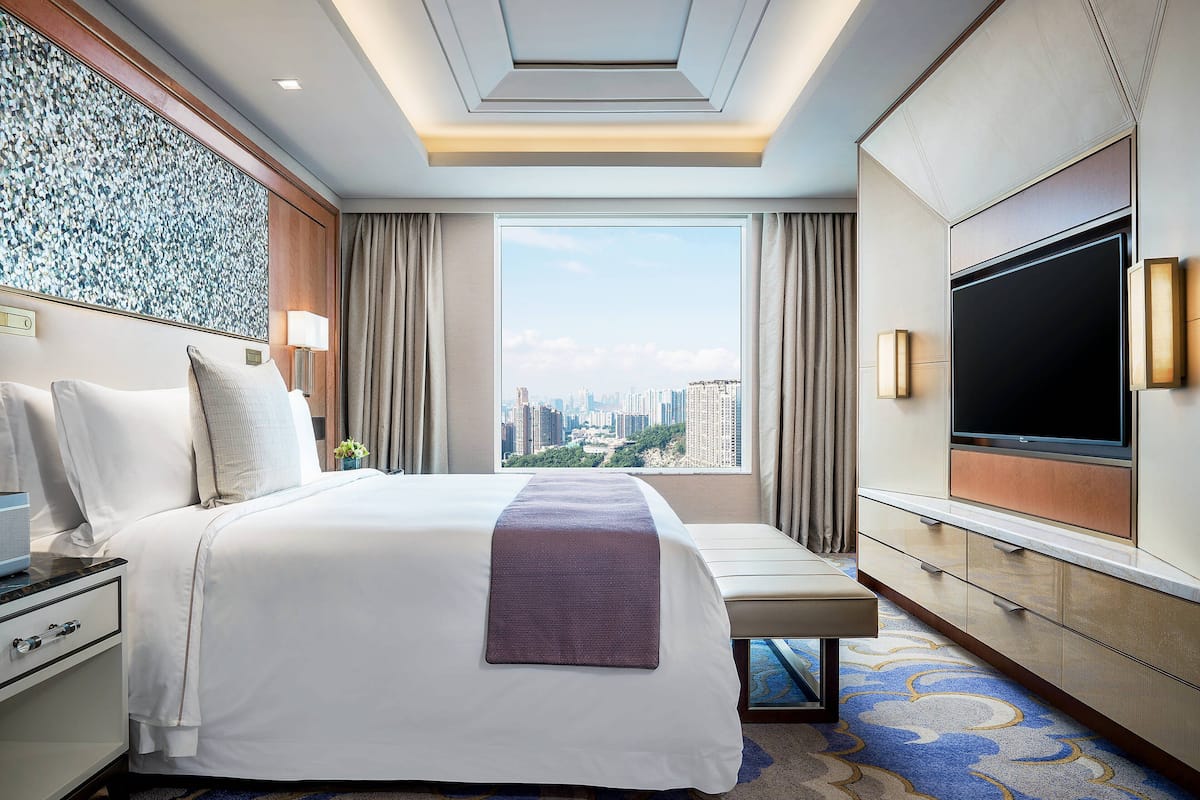 A hotel room with a large bed, a bench, a TV, and a city view.