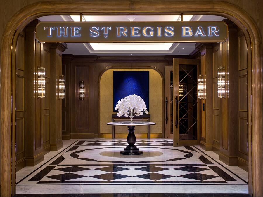 Eine Lobby mit einem Schachbrettmuster-Boden, einem Tisch mit Blumen in einer Vase und einem Schild mit der Aufschrift 'The St Regis Bar'.