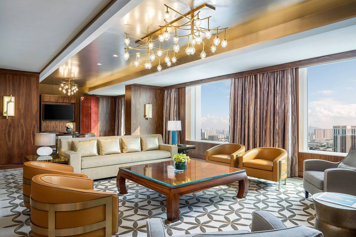 A spacious hotel room with a large window offering a city view, a modern chandelier, and a comfortable seating arrangement.