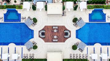 An aerial view of a hotel with two large swimming pools, a central seating area, and multiple lounge chairs around the pools.