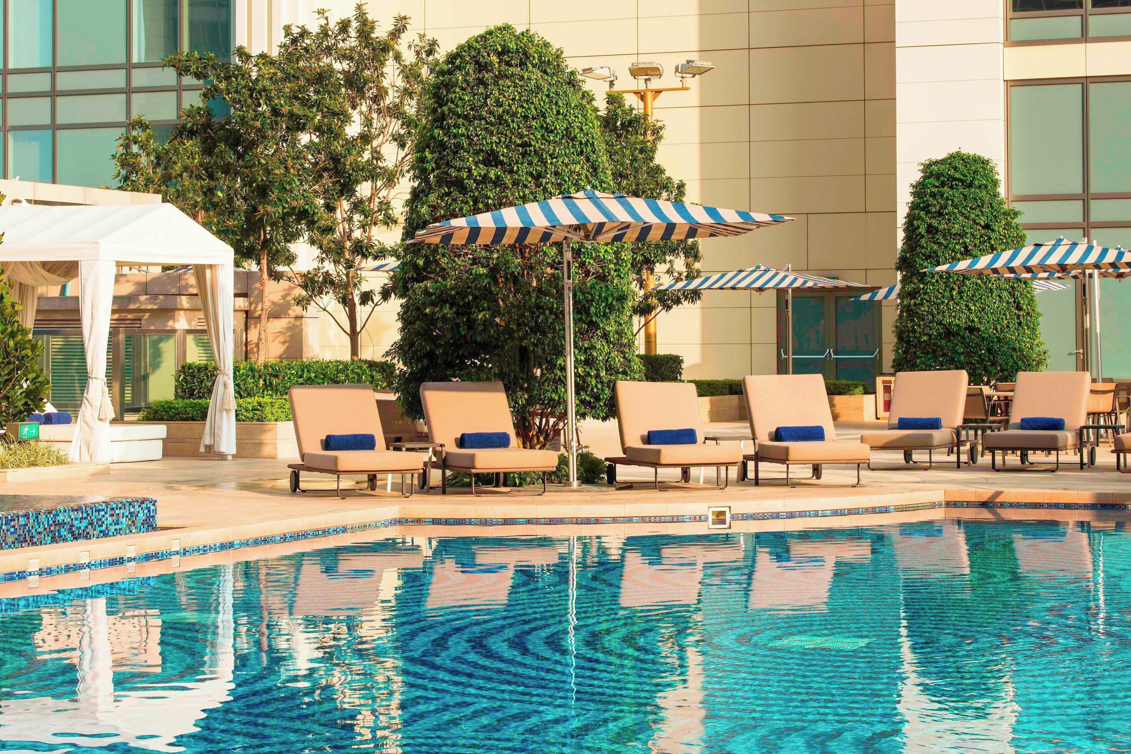 A pool area with lounge chairs and umbrellas, surrounded by greenery and a building facade.