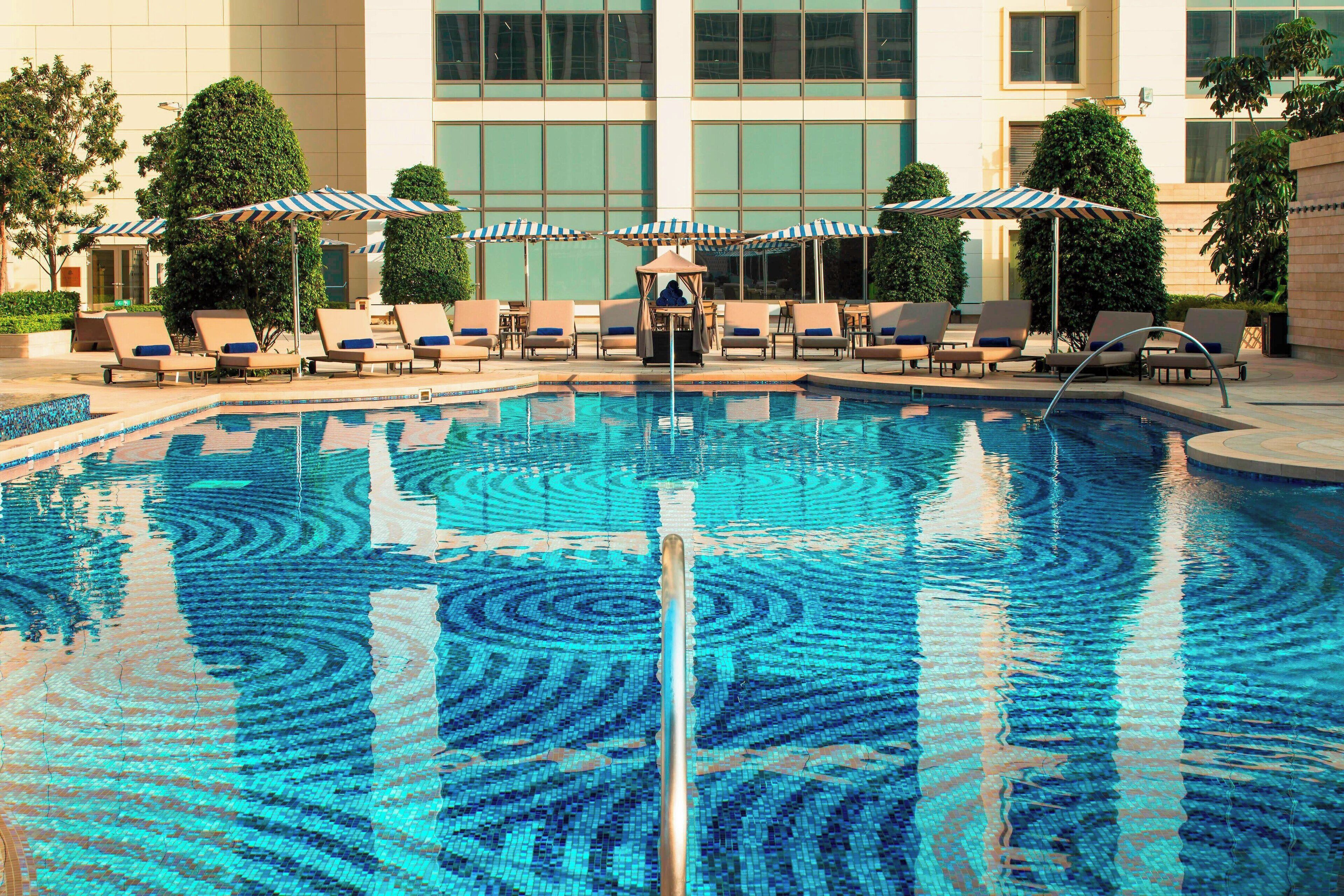 Una piscina con sillas de descanso y sombrillas, rodeada por un edificio con grandes ventanales.