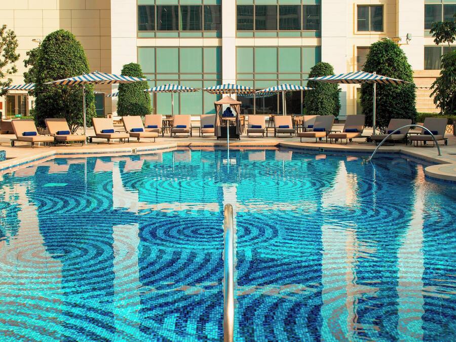 Una piscina con sillas de descanso y sombrillas, rodeada por un edificio con grandes ventanales.
